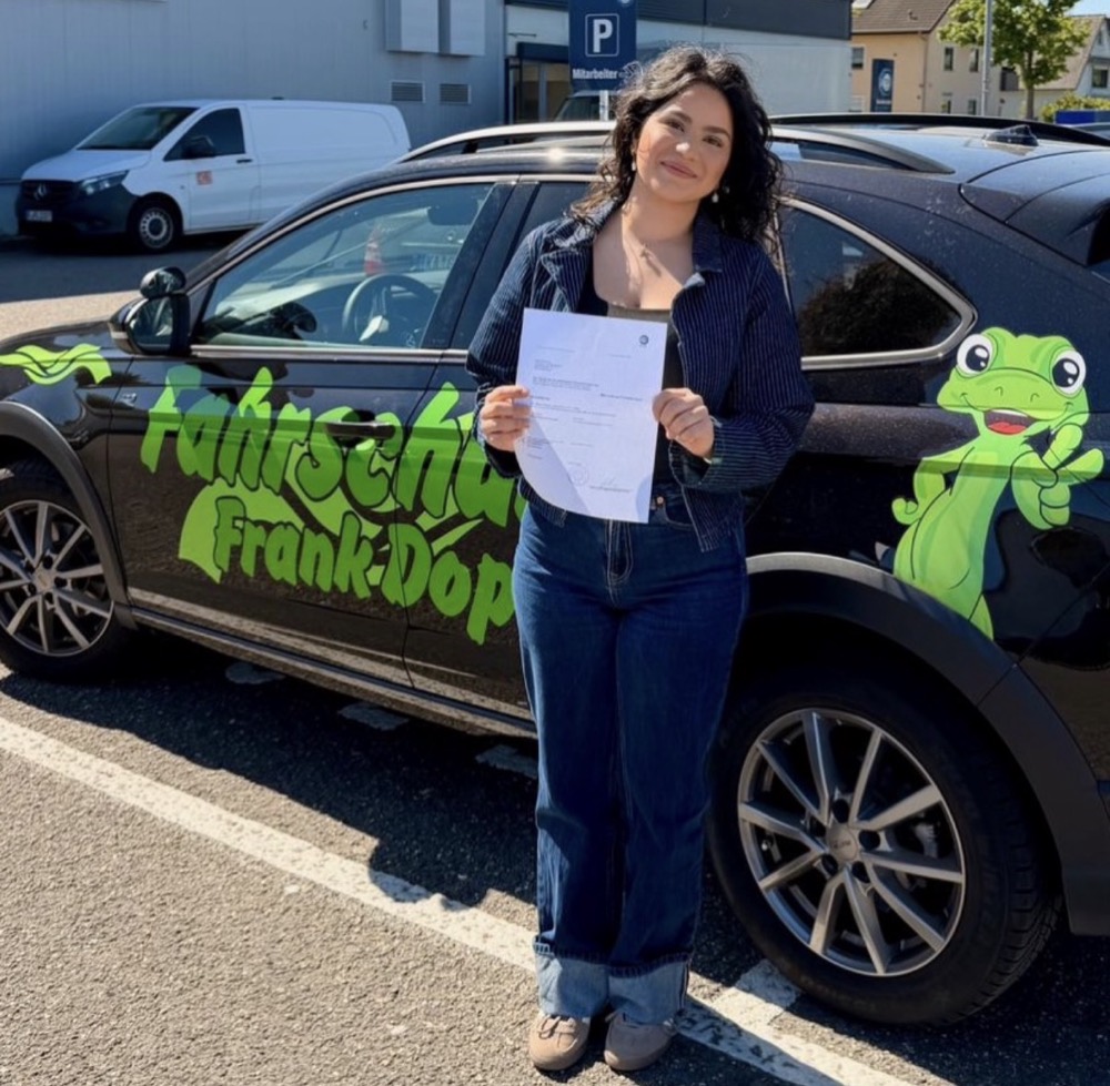 Fahrschülerin mit bestandener Prüfung neben dem Gecko-Fahrschulauto